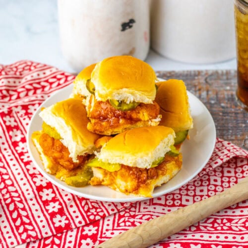 A plate of chicken sliders with pickles on a patterned red cloth, accompanied by a bowl of pickles, a wooden spoon, and a glass of iced drink.