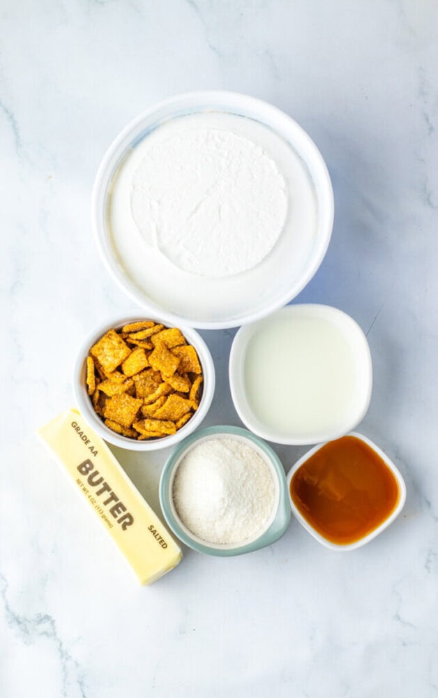 Five ingredients—heavy cream, milk, caramel sauce, shredded coconut, Cinnamon Toast Crunch cereal, and a stick of butter—arranged in small bowls on a white marble surface.
