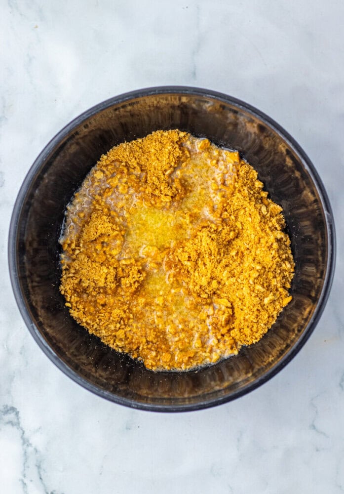 A black bowl contains a mixture of crushed graham crackers, melted butter, and sugar on a white marble surface.