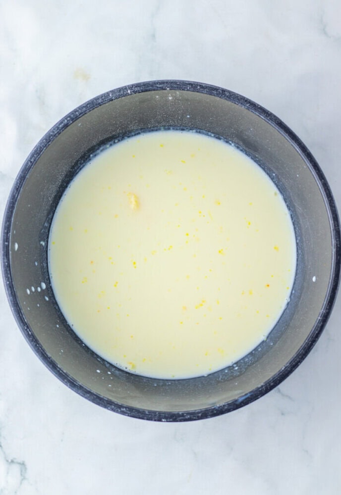 A black bowl containing a pale yellow liquid mixture with small yellow specks, placed on a white marble surface.