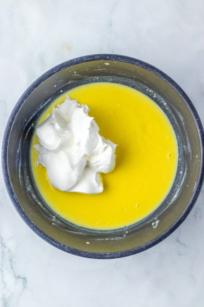 A mixing bowl containing yellow liquid mixture with a dollop of whipped cream or beaten egg whites on top, placed on a marble surface.