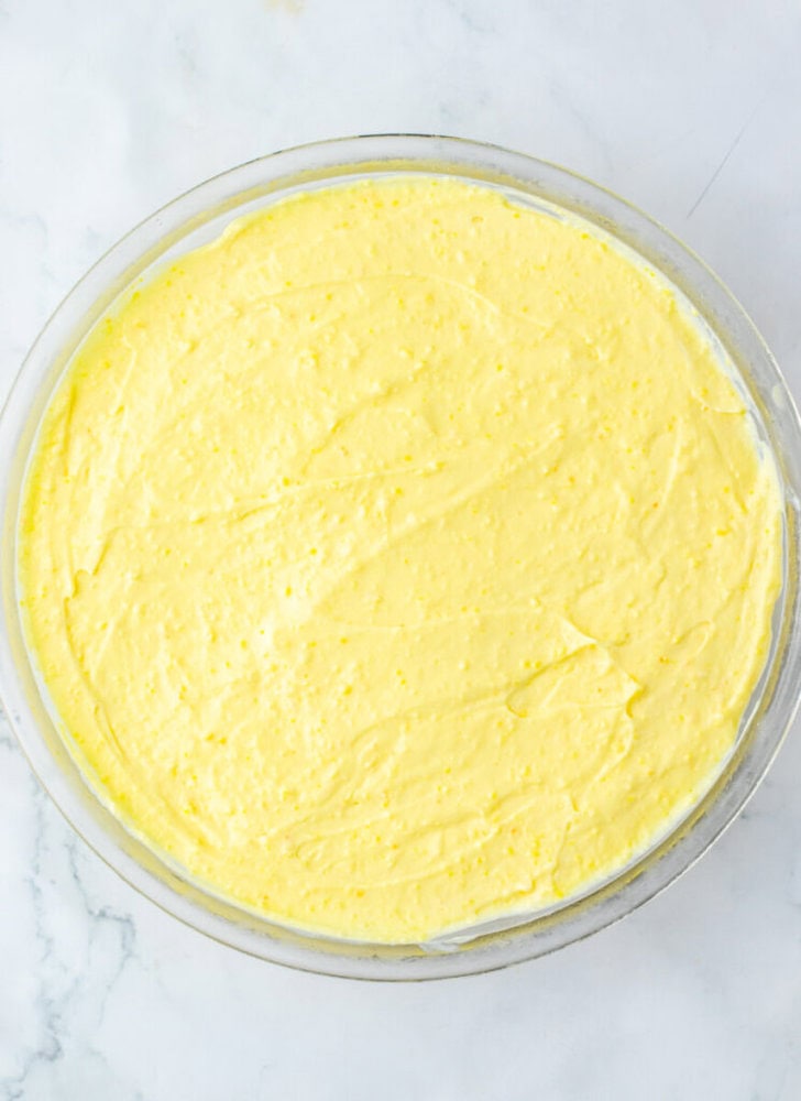 A glass bowl filled with a smooth, yellow mixture sits on a white marble surface.
