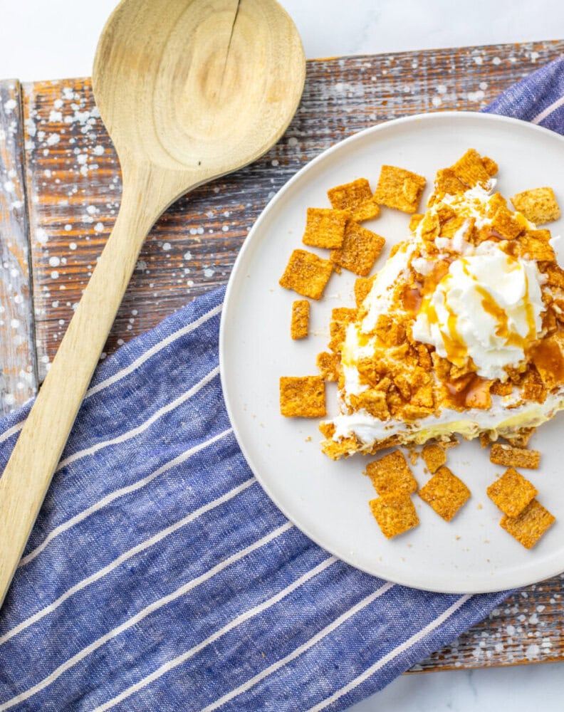 A plate with a dessert topped with whipped cream, caramel sauce, and crunchy cereal, next to a wooden spoon and a blue-striped napkin on a rustic wooden surface.