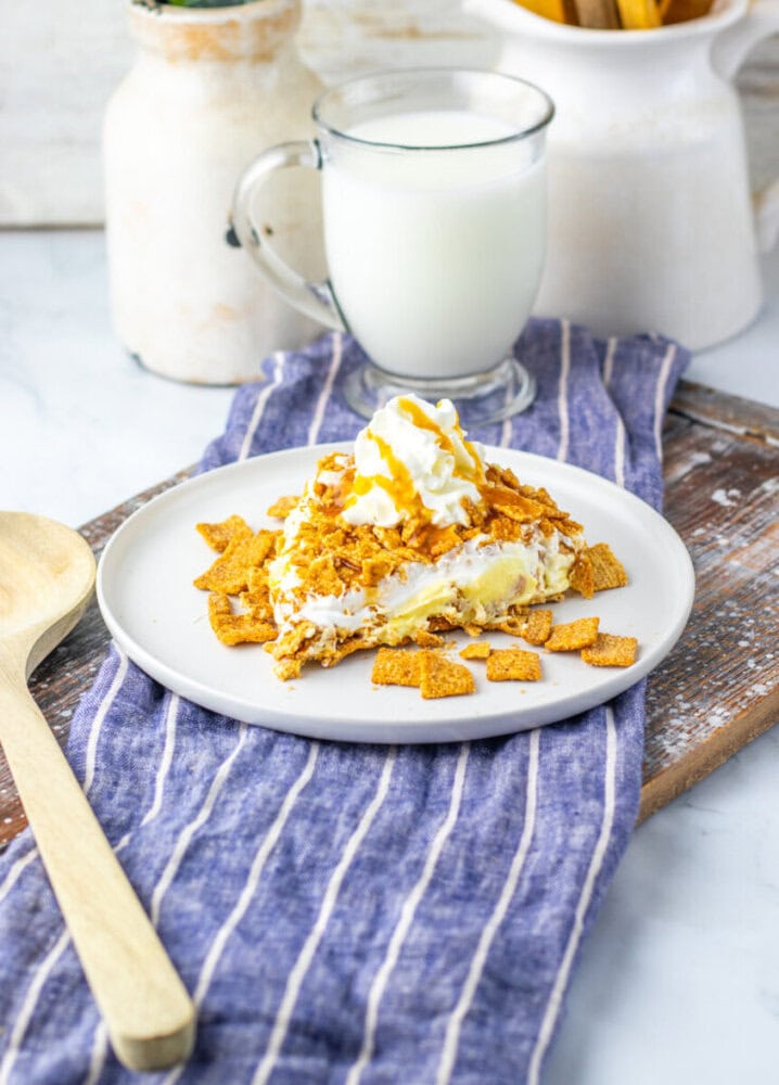 A slice of dessert topped with whipped cream and caramel sauce on a white plate, garnished with cereal pieces, next to a glass of milk on a striped cloth.