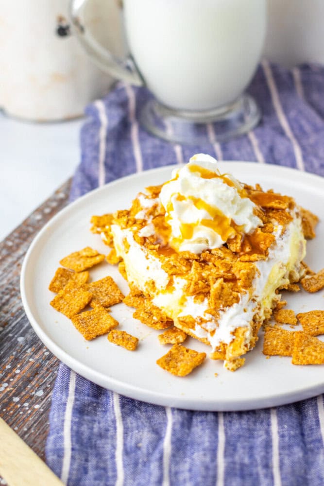 A dessert topped with cereal flakes, whipped cream, and caramel sauce on a white plate, with a glass of milk in the background.