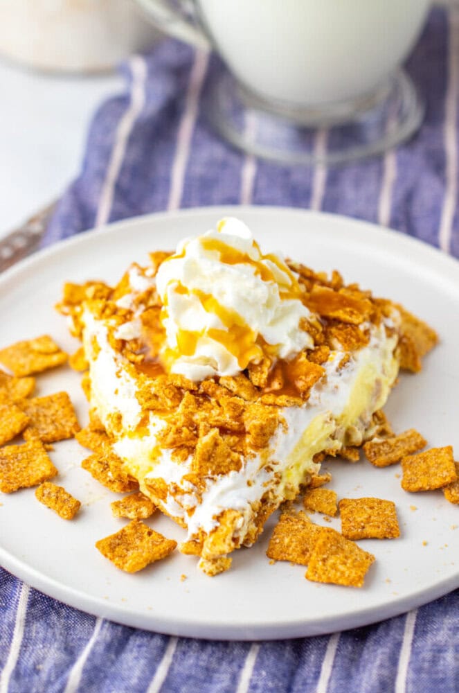 A slice of dessert topped with whipped cream and caramel sauce, with pieces of cinnamon cereal scattered on a white plate.