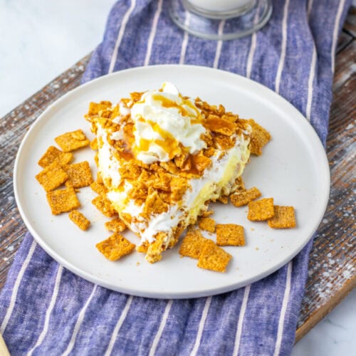 A slice of ice cream dessert topped with whipped cream, caramel drizzle, and crushed cereal pieces on a white plate, next to a wooden spoon on a striped cloth.