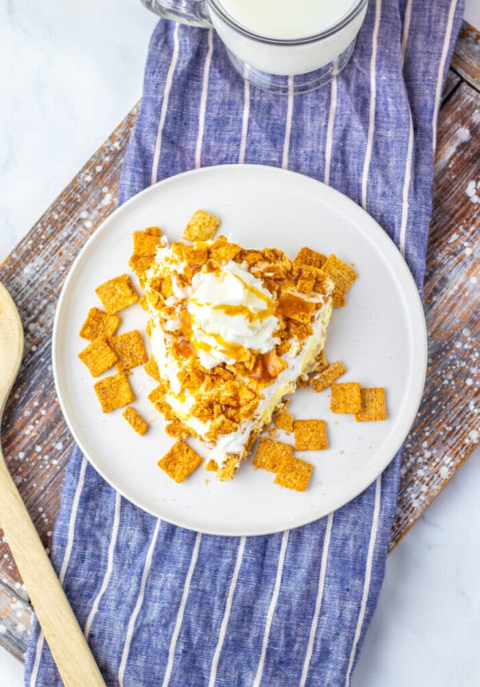 A slice of dessert topped with whipped cream and cinnamon cereal pieces on a white plate, placed on a blue-striped cloth with a wooden spoon and a glass of milk nearby.
