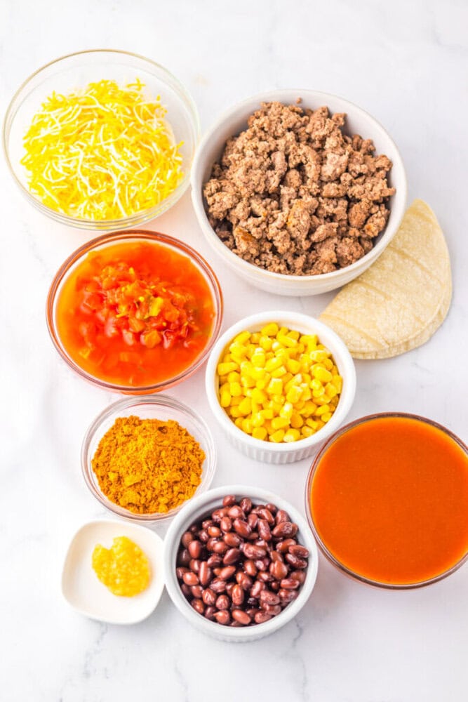 Bowls containing shredded cheese, cooked ground beef, diced tomatoes, corn, taco seasoning, beans, minced garlic, enchilada sauce, and corn tortillas are arranged on a white surface.