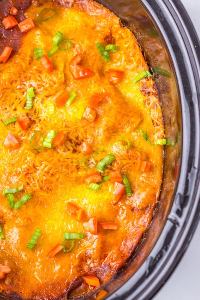 Casserole topped with melted cheese, diced tomatoes, and chopped green onions in a slow cooker.
