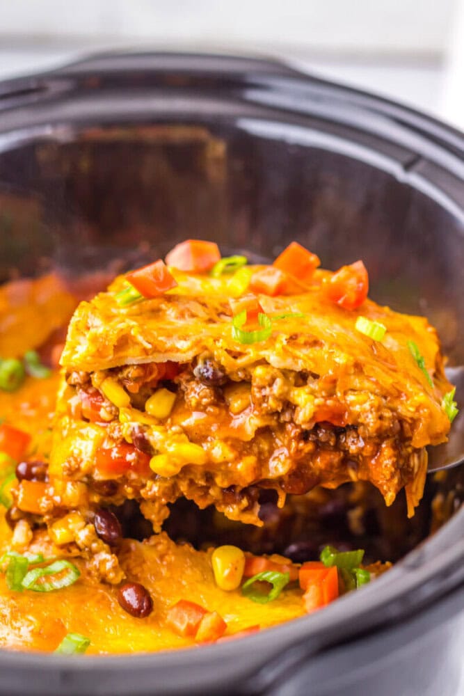 A close-up of a serving of cheesy taco casserole with ground meat, beans, corn, and diced tomatoes being lifted from a slow cooker.