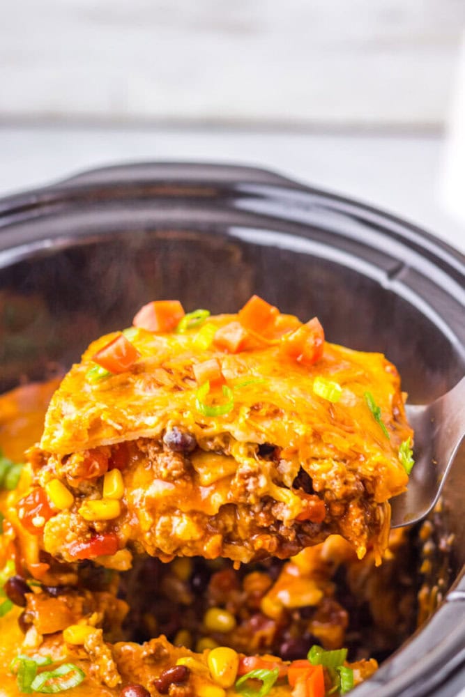 A close-up of a serving of cheesy taco casserole with ground beef, corn, black beans, and diced tomatoes being lifted from a slow cooker.