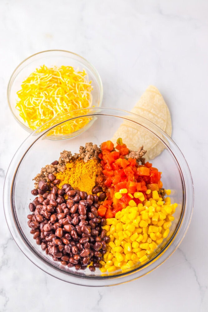 A glass bowl with black beans, corn, diced tomatoes, cooked ground meat, and seasoning. Beside it are corn tortillas and a bowl of shredded cheese.