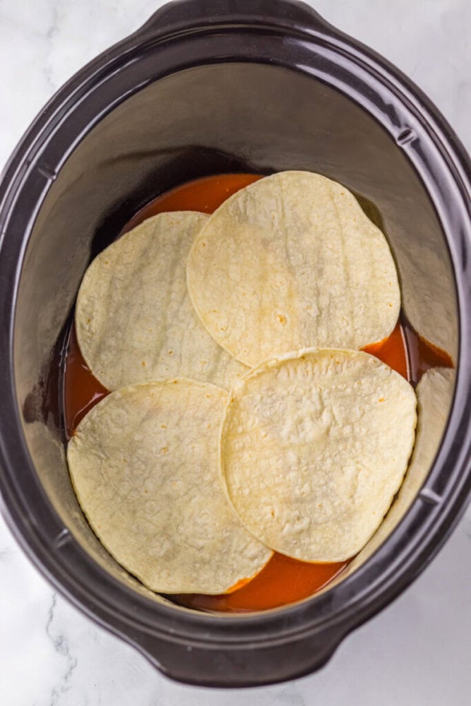 Four corn tortillas are layered on top of red sauce inside a slow cooker.