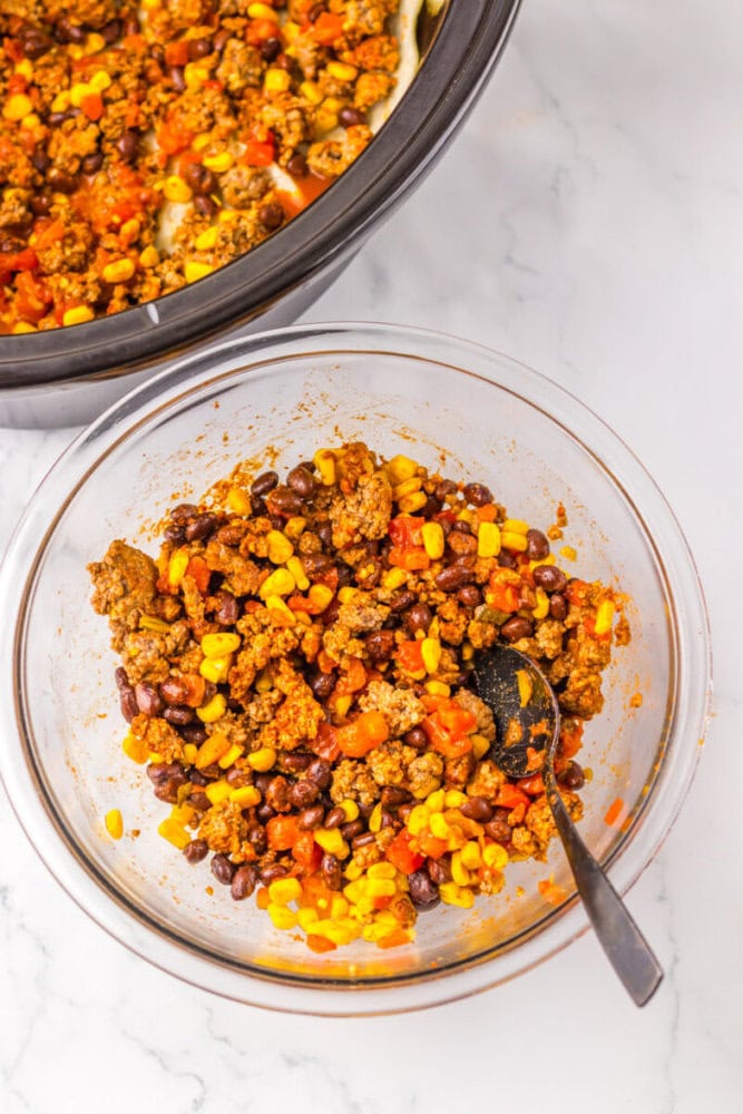 A glass bowl filled with a mixture of ground meat, corn, black beans, and diced tomatoes, with a black spoon inside; a slow cooker is partially visible above.