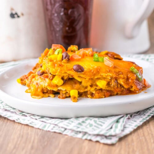 A slice of casserole with cheese, black beans, corn, and vegetables on a white plate, placed on a patterned napkin with a drink in the background.