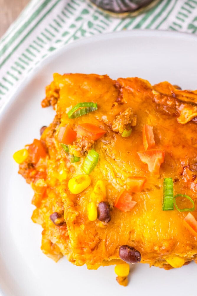 A close-up of a serving of cheesy casserole with ground meat, black beans, corn, tomatoes, and green onions on a white plate.