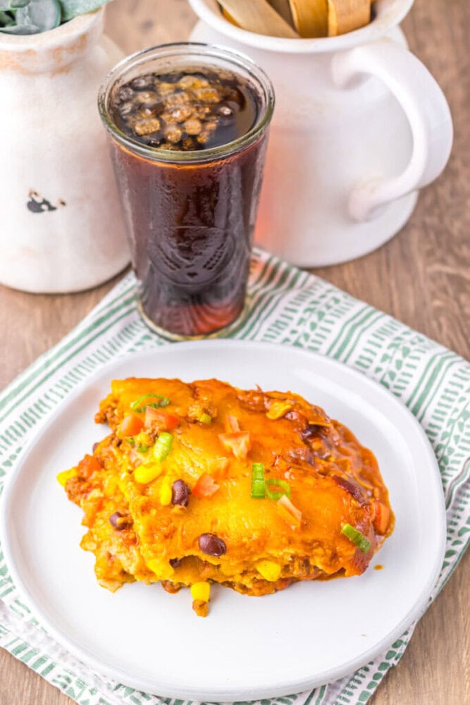 A serving of cheesy casserole with corn, beans, and green onions on a white plate, next to a glass of dark soda on a green patterned napkin.