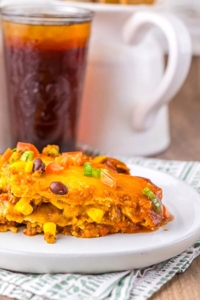 A serving of cheesy casserole with beans, corn, and ground meat on a white plate, accompanied by a glass of dark soda in the background.
