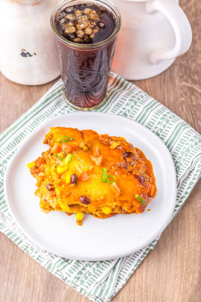 A serving of cheesy casserole with beans and corn on a white plate, next to a glass of dark soda on a green patterned napkin.