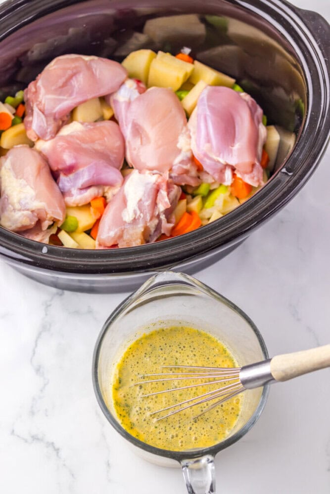 Uncooked chicken thighs on top of chopped vegetables in a slow cooker, with a measuring cup of seasoned liquid and a whisk beside it on a marble countertop.