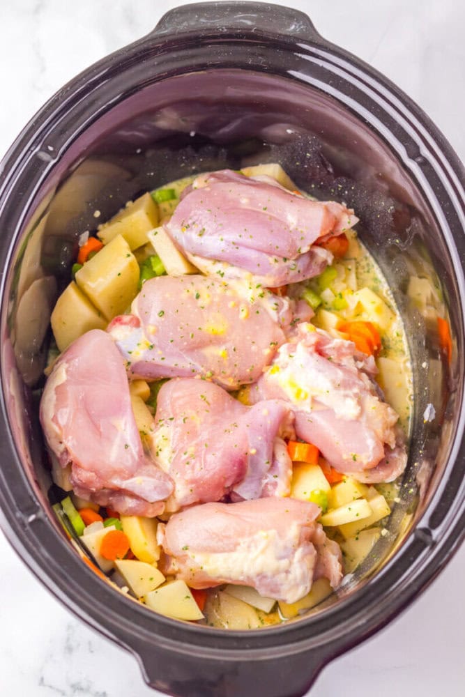 Raw chicken pieces seasoned with herbs are placed on top of chopped potatoes, carrots, and celery inside a slow cooker.