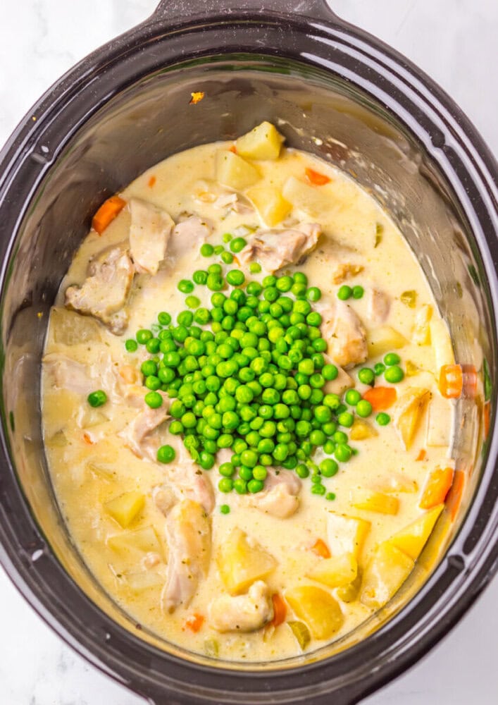 Chicken stew with potatoes, carrots, and a creamy sauce in a slow cooker, topped with a pile of green peas before mixing.