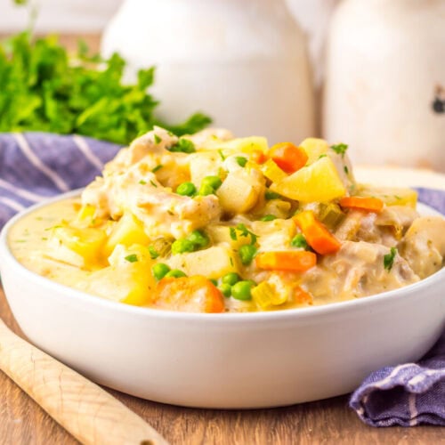 A bowl of creamy chicken pot pie filling with peas, carrots, potatoes, and chicken sits on a wooden table next to a wooden spoon and a blue-striped cloth.