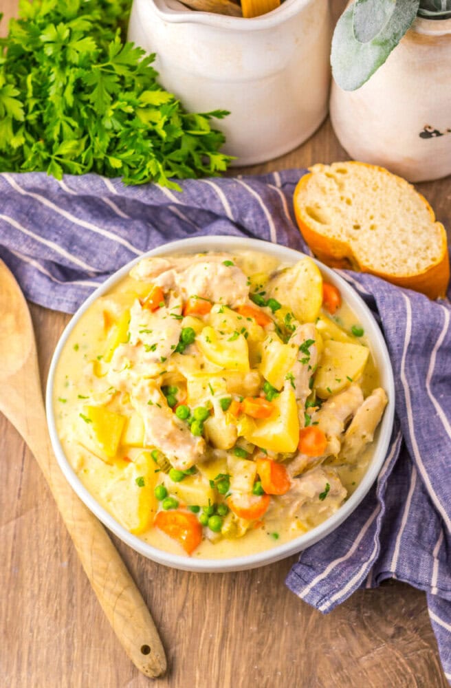 A bowl of creamy chicken stew with potatoes, carrots, peas, and herbs on a wooden table, next to bread, parsley, and kitchen utensils.