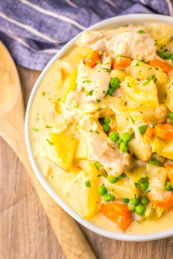 A bowl of creamy chicken stew with peas, carrots, potatoes, and herbs, next to a wooden spoon on a wooden table.