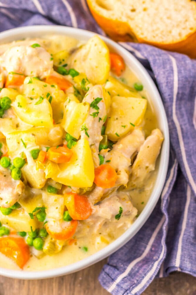 A bowl of creamy chicken stew with potatoes, carrots, and peas, garnished with herbs, next to a slice of bread on a blue-striped cloth.