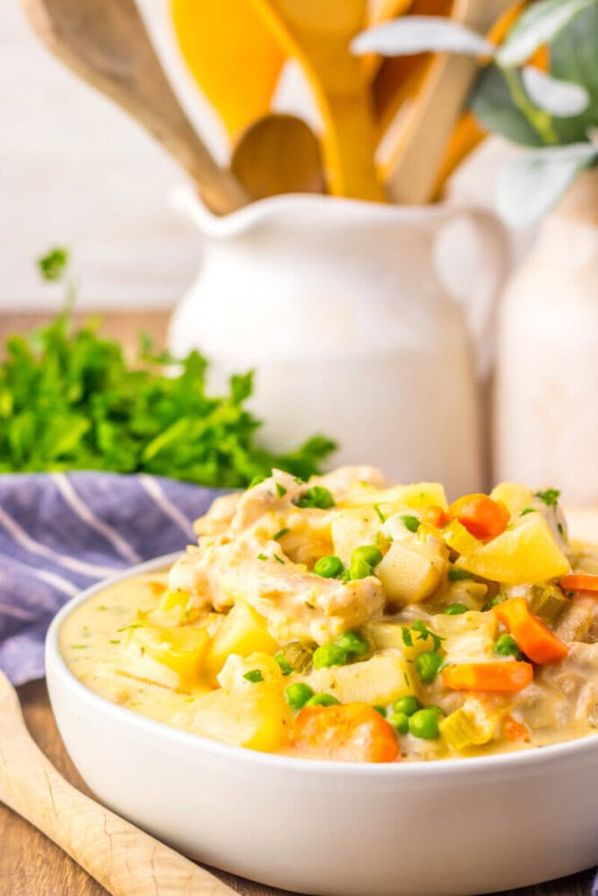 A bowl of creamy vegetable stew with carrots, potatoes, peas, and herbs sits on a wooden table with fresh parsley and kitchen utensils in the background.