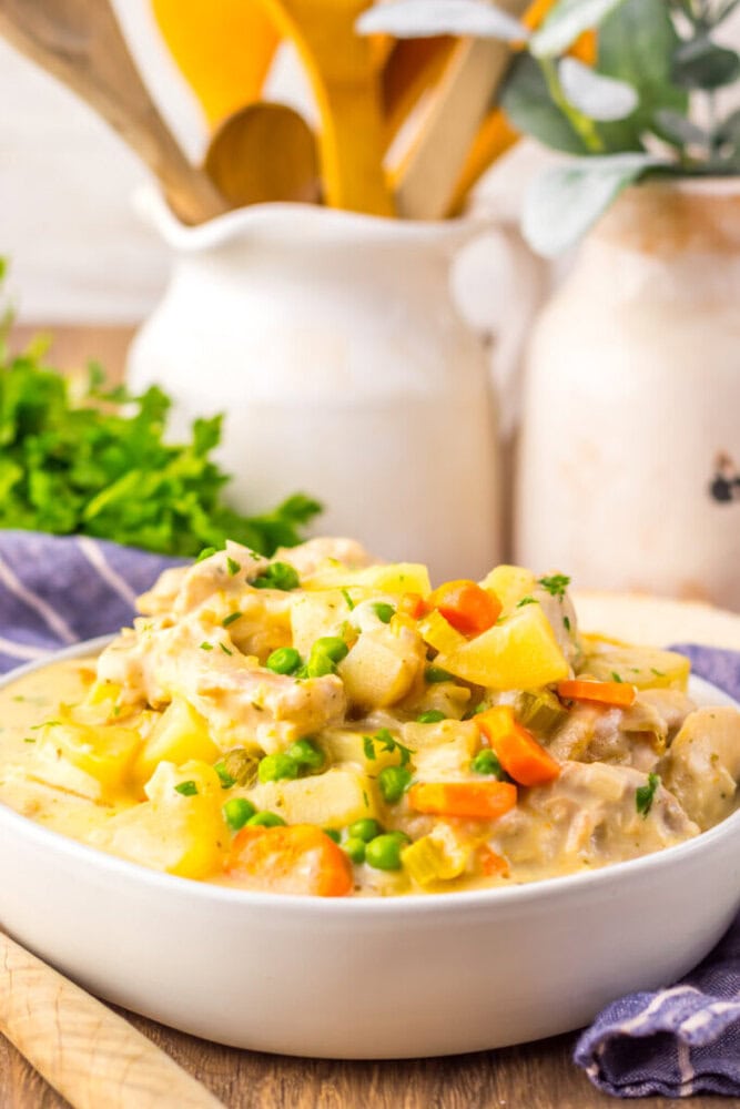 A bowl of creamy chicken stew with potatoes, carrots, peas, and herbs, placed on a table with kitchen utensils and greenery in the background.