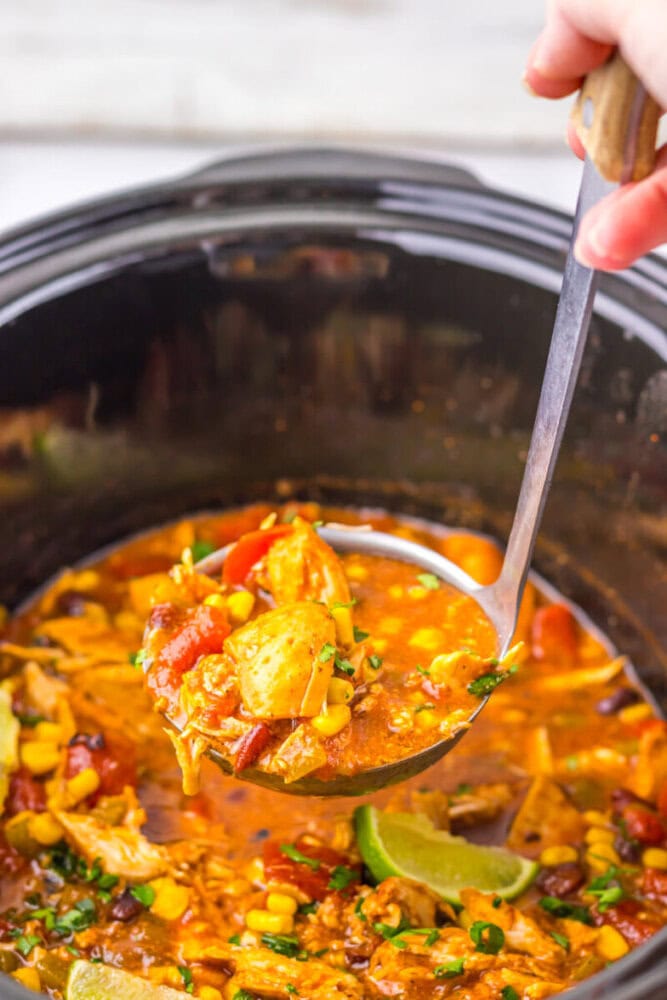 A hand holds a ladle of chunky chicken soup with corn, tomatoes, and vegetables over a slow cooker filled with the same soup.
