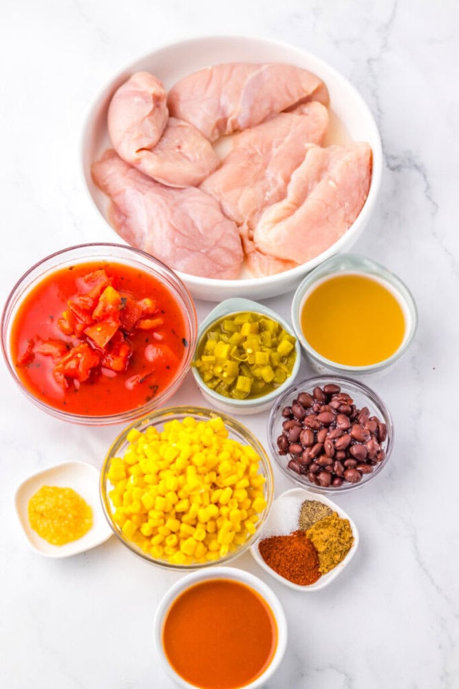 Raw chicken breast, diced tomatoes, chopped green chiles, corn, black beans, broth, garlic, hot sauce, and spices arranged in bowls on a white surface.