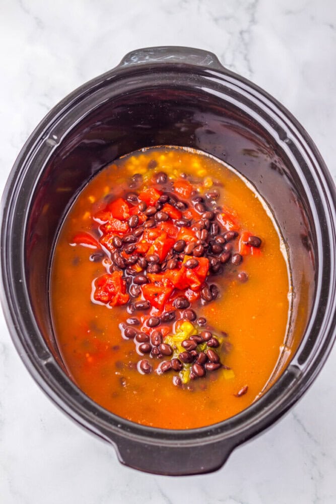 A slow cooker filled with black beans, diced tomatoes, broth, and vegetables on a marble countertop.