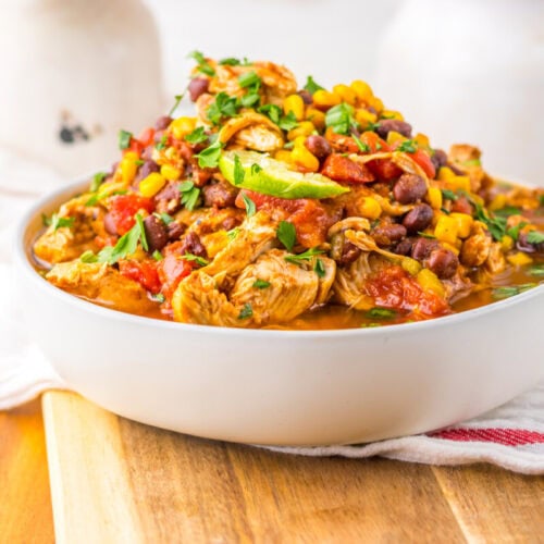 A bowl of chicken chili with black beans, corn, tomatoes, and herbs sits on a wooden board with a white towel in the background.