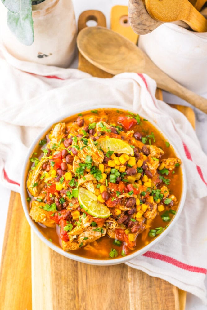 A bowl of chicken chili with black beans, corn, tomatoes, and lime wedges, garnished with chopped herbs, sits on a wooden board with kitchen utensils in the background.