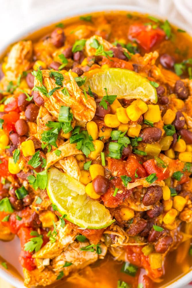 A close-up of a dish featuring shredded chicken, black beans, corn, tomatoes, and lime wedges, garnished with chopped green onions and cilantro.