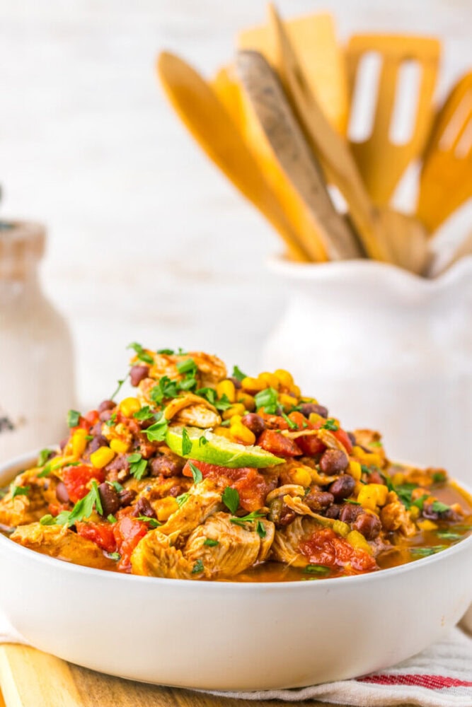 A bowl of chili with shredded chicken, black beans, corn, tomatoes, and herbs sits on a table with wooden utensils in the background.