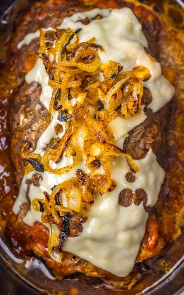A cooked meatloaf topped with melted cheese slices and caramelized onions, sitting in a pool of its own juices.