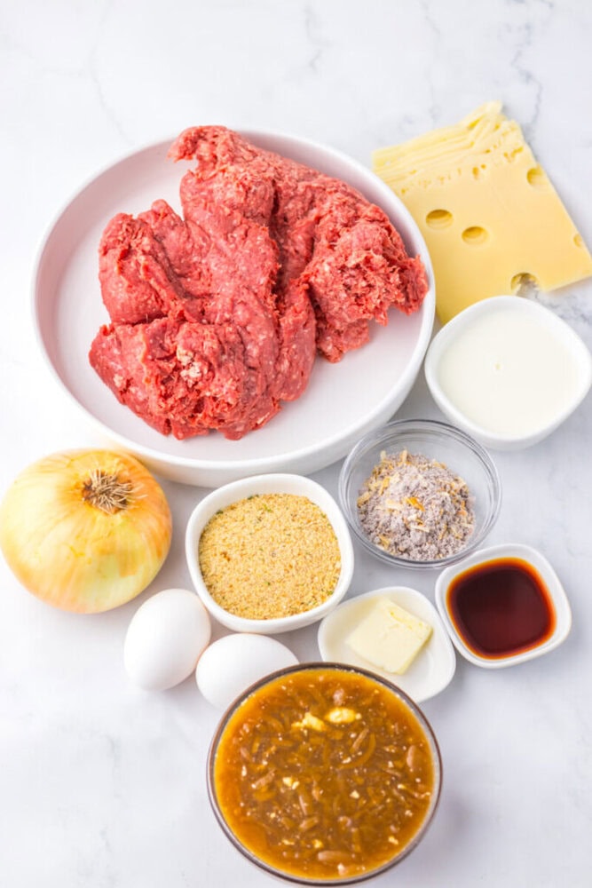 Raw ground beef on a plate surrounded by slices of Swiss cheese, an onion, eggs, bread crumbs, milk, butter, Worcestershire sauce, and a bowl of soup mixture on a white surface.