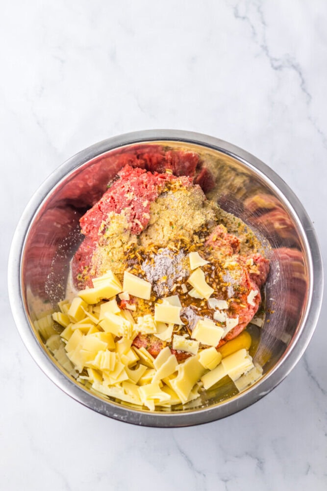 A metal mixing bowl filled with ground meat, shredded cheese, breadcrumbs, chopped onions, and various seasonings on a white marble surface.