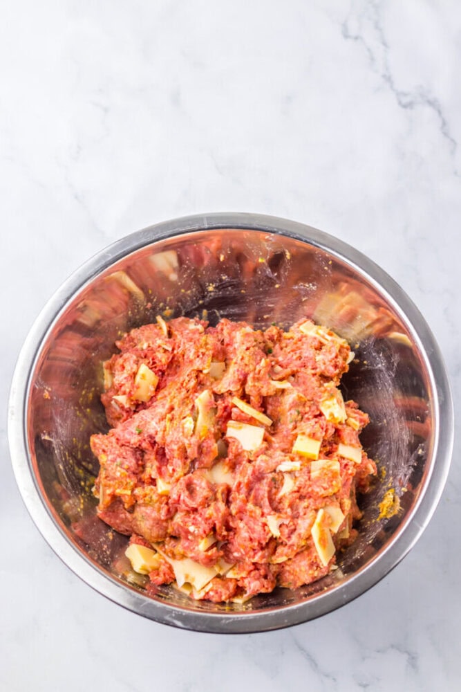 A stainless steel bowl filled with a mixture of ground meat and cheese chunks on a white marble surface.