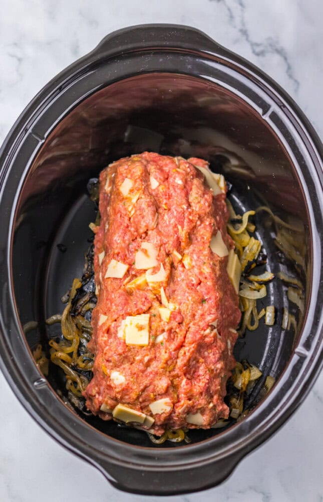 Raw meatloaf mixture with cheese on top of cooked onions inside a slow cooker, ready to be cooked.