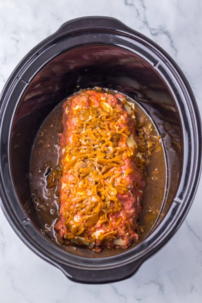 A meatloaf topped with cooked onions and sauce sits in a slow cooker with liquid at the bottom.