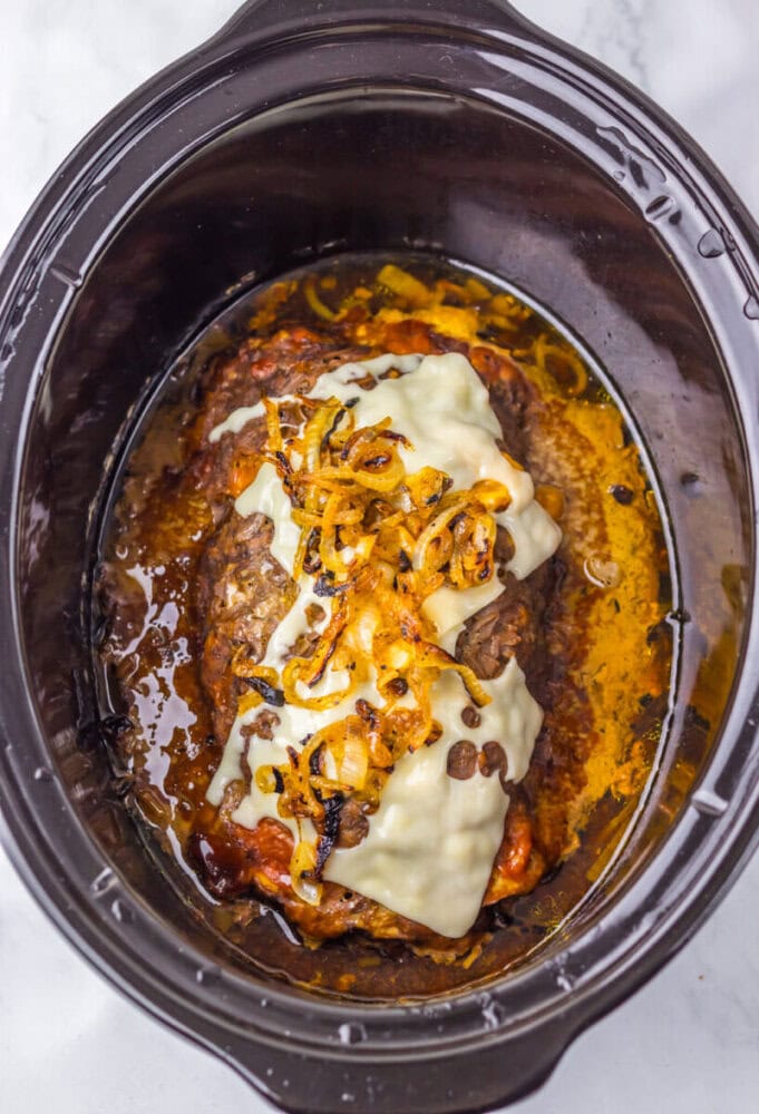 A cooked meatloaf topped with melted cheese and sautéed onions inside a black slow cooker.