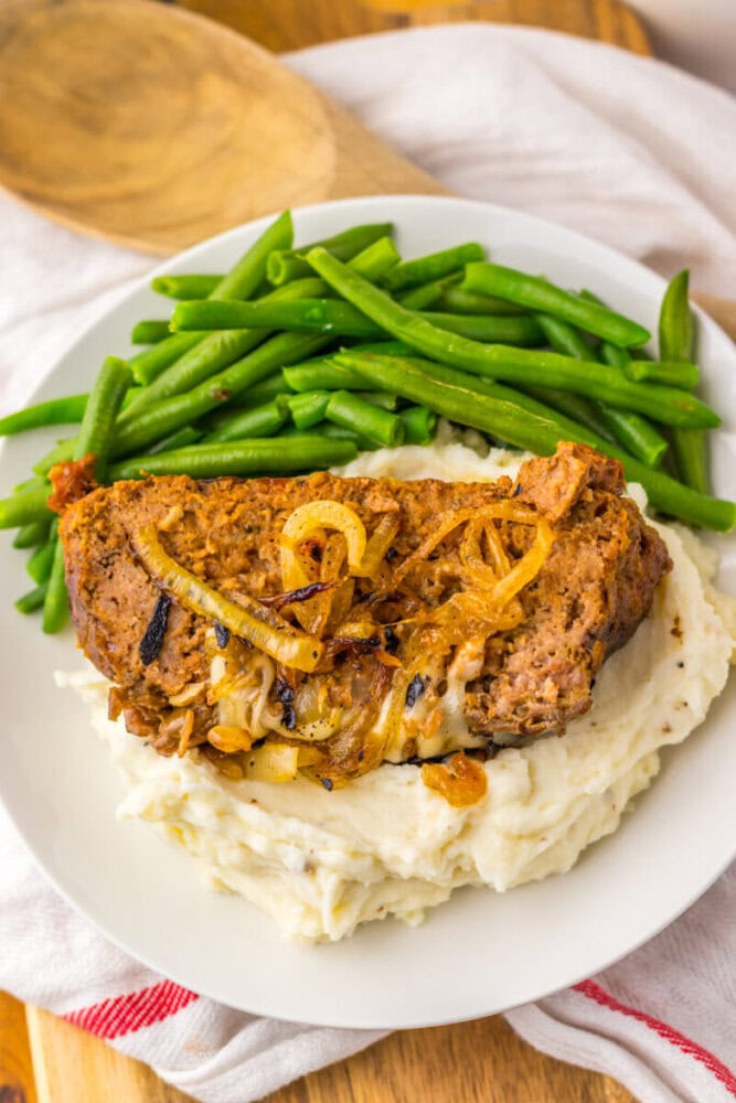 A plate with a slice of meatloaf topped with onions, served on mashed potatoes with a side of green beans.