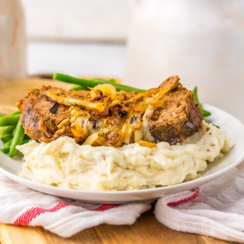 A plate of mashed potatoes topped with two slices of meatloaf and sautéed onions, served with green beans.