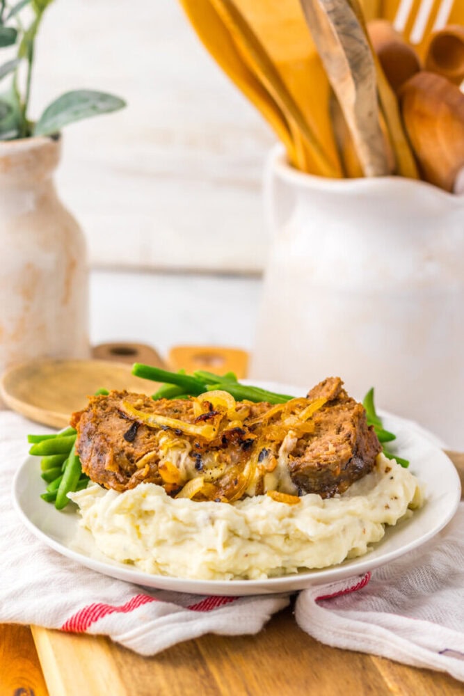 A plate of meatloaf topped with caramelized onions, served on mashed potatoes with green beans, sits on a white plate with kitchen utensils in the background.